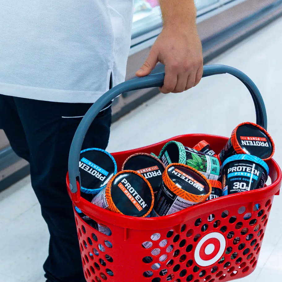 Person holding a red Target shopping basket filled with protein powder containers in a store.

